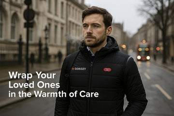 Man wearing a black GOKOZY men's heated jacket with hood walking a UK city street with red bus and Georgian buildings, headline text 'Wrap Your Loved Ones in the Warmth of Care'